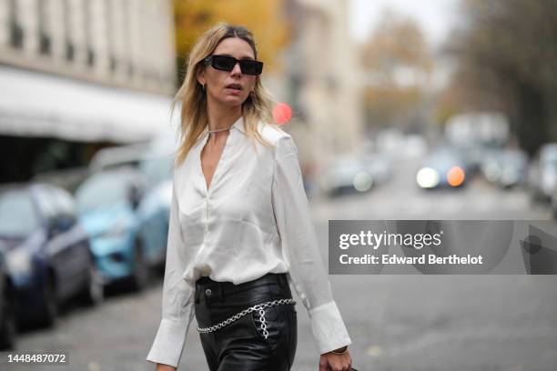 Natalia Verza wears black squared sunglasses from Prada, silver earrings, a white silk buttoned shirt, black shiny leather large wide legs pants, a...