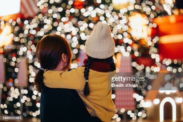 rear view of young asian mother carrying her lovely little daughter in arms, looking at illuminated christmas tree in christmas market. family moment. enjoying christmas atmosphere. holiday and festive vibes. christmas is here - christmas celebrations in china imagens e fotografias de stock