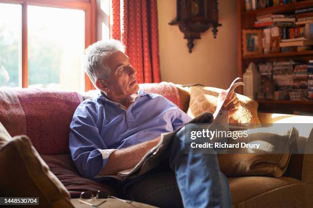 portrait of a senior man sitting on the sofa reading a newspaper - newspapers uk stock-fotos und bilder