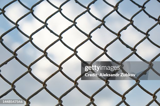 Metal Wire Mesh On A Background Of Blue Sky With White Clouds. Fence, barbed wire. The Concept Of Restriction Of Freedom. Sanctions against the country. Problems, economic and financial crisis due to military actions and offenses. Metal Wire Mesh On A Background Of Blue Sky With White Clouds. Fence, barbed wire. The Concept Of Restriction Of Freedom. Sanctions against the country. Problems, economic and financial crisis due to military actions and offenses.