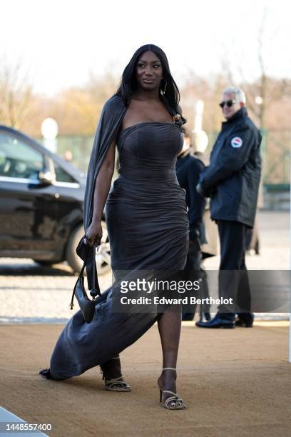 Aya Nakamura wears a gray low-neck and sleeveless see-through maxi dress, a black leather bag, high heeled shoes, outside the "Le Raphia" Jacquemus...