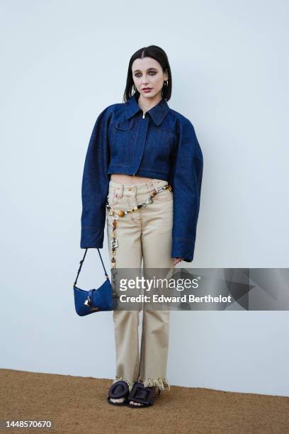 Emma Chamberlain wears a blue denim cropped oversized jacket with long sleeves, beige pants with attached long jewelry, a blue suede Jacquemus bag...