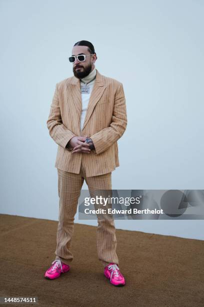 Wears sunglasses, a metallic silver necklace with incsriptions, a white turtleneck pullover, a beige striped blazer jacket, matching suit pants, pink...