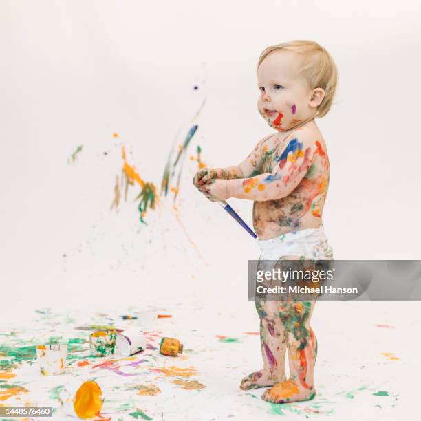 a young toddler, covered in paint, plays with washable paint on a white backdrop. - baby paint stock pictures, royalty-free photos & images