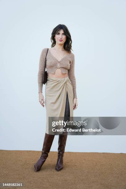 Jeanne Damas wears a pale brown wool cropped cardigan, a beige slit midi skirt, black tights, a bag, brown pointed knee-high leather boots, outside...