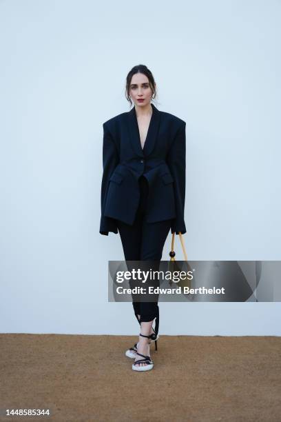 Rebecca Dayan wears a black oversized blazer jacket, black leggings, a yellow bag, white high heeled lace-up shoes, outside the "Le Raphia" Jacquemus...