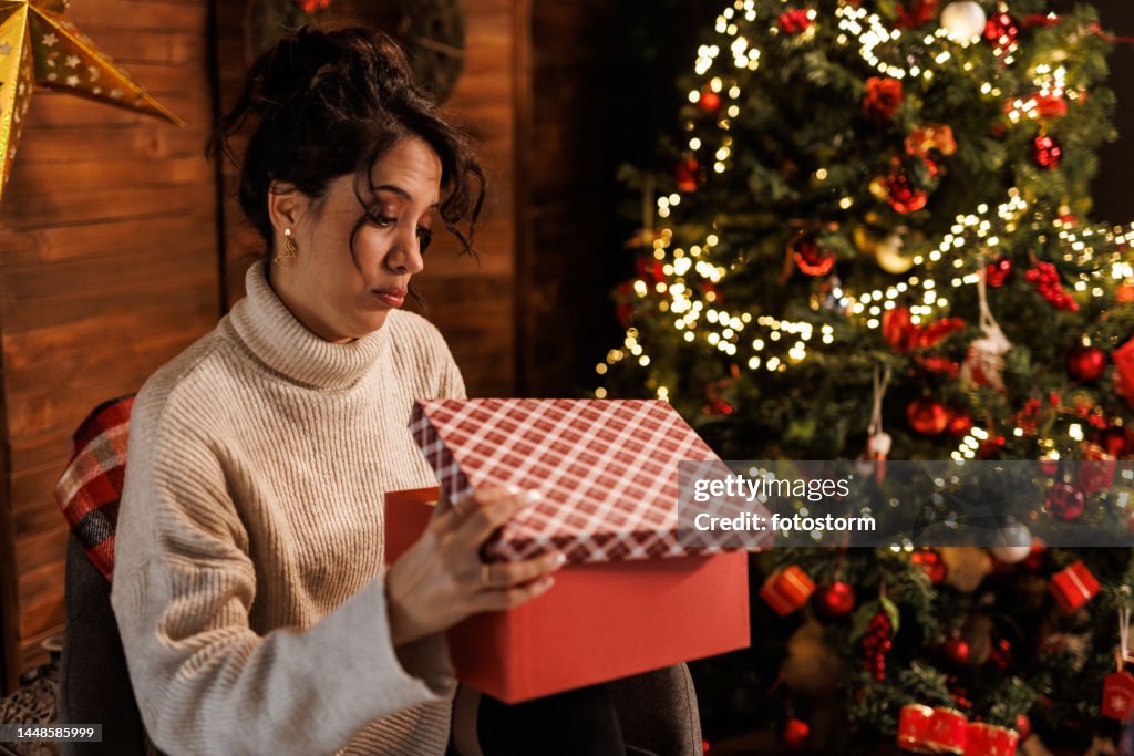 Giovane donna che si siede vicino all'albero di Natale, aprendo una scatola regalo