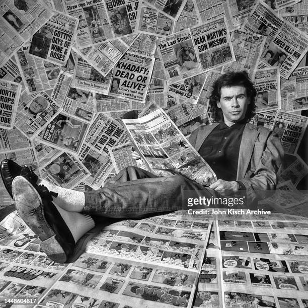 Portrait of American comedian and talk-show host Dennis Miller as he sits, feet up on a table covered in newspapers, New York, New York, 1987.