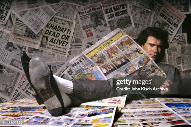 Portrait of American comedian and talk-show host Dennis Miller as he sits, feet up on a table covered in newspapers, New York, New York, 1987.