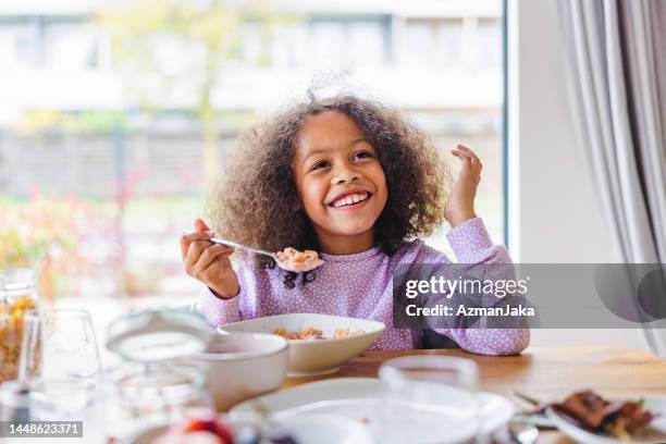 happy adorable black little girl smiling while eating cereal for breakfast in a beautiful kitchen - children eating breakfast stock pictures, royalty-free photos & images