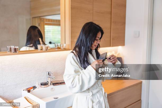 attractive brunette female in a bathrobe carefully brushing her hair while leaning on the bathroom sink in a beautiful bathroom - woman brushing hair stockfoto's en -beelden