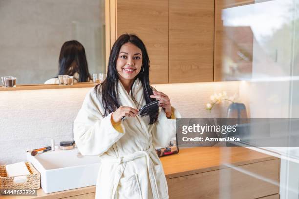 happy caucasian female in a bathrobe leaning on the bathroom sink and brushing her hair in a luxurious bathroom while looking at the camera - brushing hair stock pictures, royalty-free photos & images