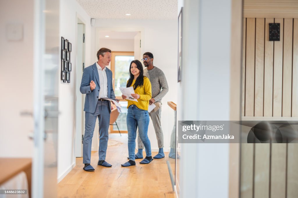Erwachsener kaukasischer männlicher Immobilienmakler, der die moderne Wohnung verkauft und seinen verschiedenen jungen Kunden zeigt, die neben ihm in der schicken Wohnung stehen