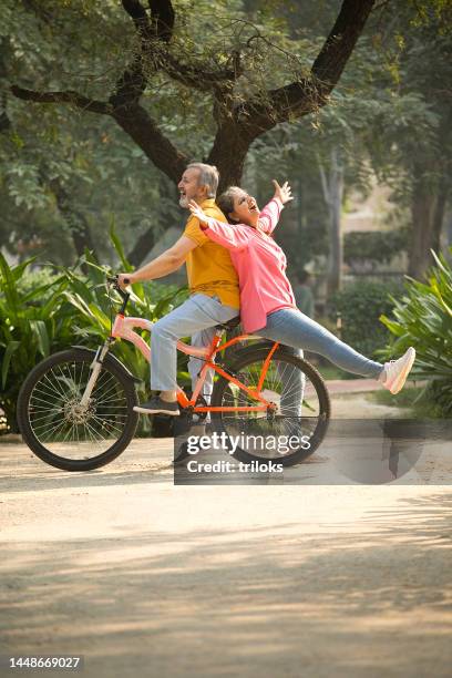 carefree wife sitting behind husband riding bicycle in park - indian elderly couple stock pictures, royalty-free photos & images