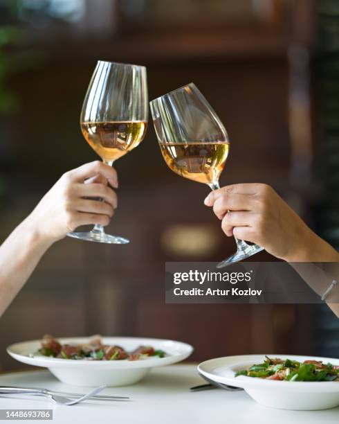 two women are drinking wine. two glasses of white wine in female hands. lunch at restaurant. wine and vegetable salad. italian food. italian restaurant. soft focus - chardonnay grape 個照片及圖片檔