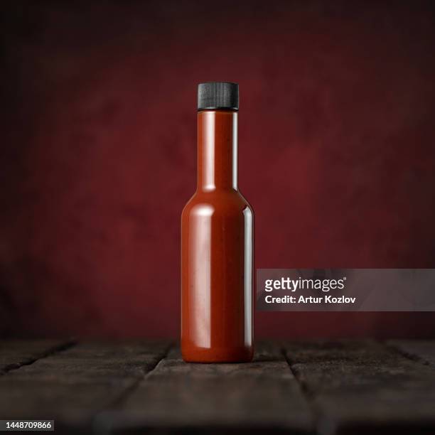 glass bottle of red hot sauce on wooden surface. chili pepper sauce. ketchup. tomato sauce. vegetarian food. photo for advertising. dark red background. copy space. soft focus. side view - hot sauce stock pictures, royalty-free photos & images