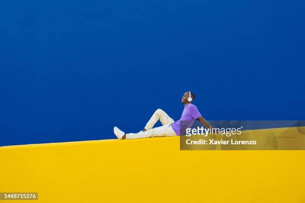 happy young man with headphones sitting on yellow wall - music inspired fashion stock pictures, royalty-free photos & images