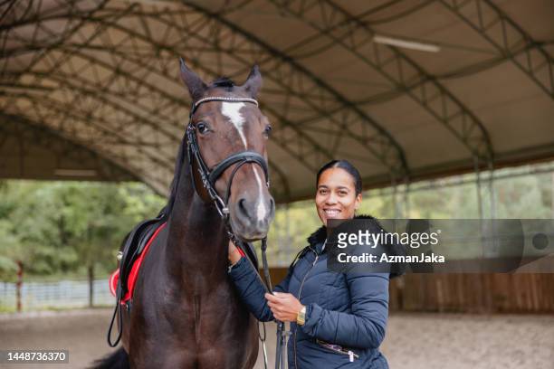 eine lächelnde schwarze junge erwachsene pferdetrainerin, die in die kamera schaut, während sie neben einem großen braunen pferd in einer reithalle auf dem land steht - sattel und zaumzeug stock-fotos und bilder