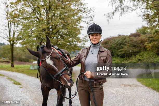 adult caucasian blonde female horse rider caressing a horse while smiling at the camera on a country road - equestrian helmet stock pictures, royalty-free photos & images