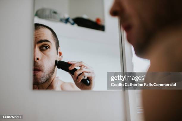 a man shaves his face with an electric shaver - standing mirror stock pictures, royalty-free photos & images