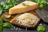Shredded parmesan cheese and grater on a cutting board. Grana padano cheese whole wedge and grated, stainless steel grater and green basil herb over wooden background. Hard cheese.