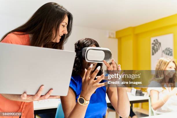 teacher in classroom holding laptop standing next to black female student with hand on her man while instructing her on how to use virtual reality goggles - interaktivität stock-fotos und bilder