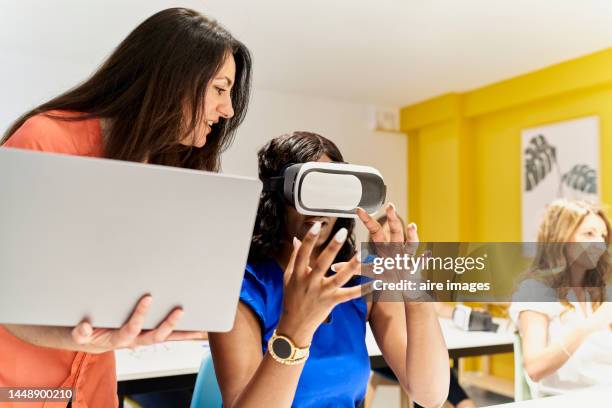 teacher in classroom holding laptop standing next to black female student with hand on her man while instructing her on how to use virtual reality goggles - virtual reality classroom stock pictures, royalty-free photos & images