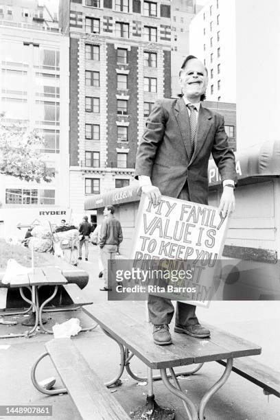 View of a demonstrator, in a suit and mask holds a sign while standing on a picnic table during a protest against Operation Rescue , organized by...