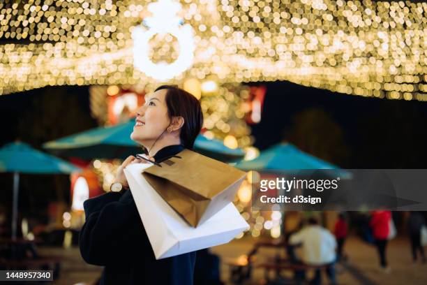 beautiful smiling young asian woman carrying paper shopping bags, shopping christmas gifts in the city. looking up at illuminated fairy lights in decorated city street. enjoying christmas shopping. festive vibes. christmas is almost here - indulgence stock pictures, royalty-free photos & images