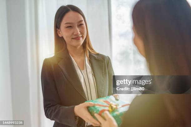 feminism woman power. two young smart beautiful asian office lady businesswomen smiling and doing gift exchange in front of a nicely decorated christmas tree in a meeting room - overdracht business mensen stockfoto's en -beelden