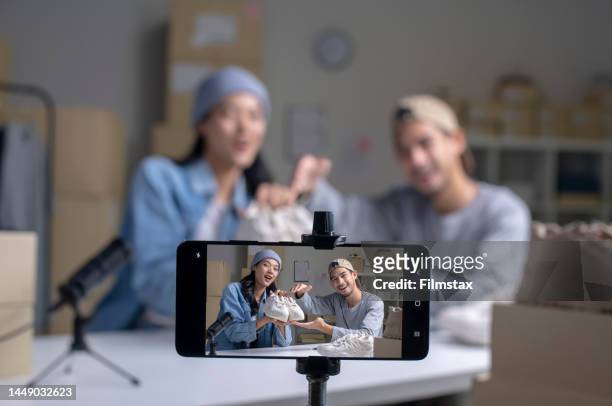 joven pareja asiática propietaria de un negocio de comercio electrónico hablando y mirando la cámara revisando el producto. - servizio fotografico fotografías e imágenes de stock
