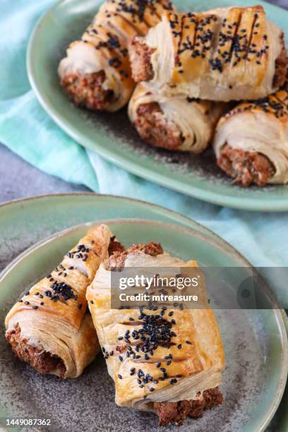 close-up image of green plates containing rows of baked, homemade sausage rolls topped with black sesame seeds, decorated with diagonal scored lines, turquoise muslin, mottled, grey background, elevated view, focus on foreground - sausage roll stock pictures, royalty-free photos & images