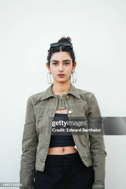 Guest wears black squared sunglasses, silver long pendant earrings, a khaki denim jacket, a black shoulder-off / cropped top, high waist black large...