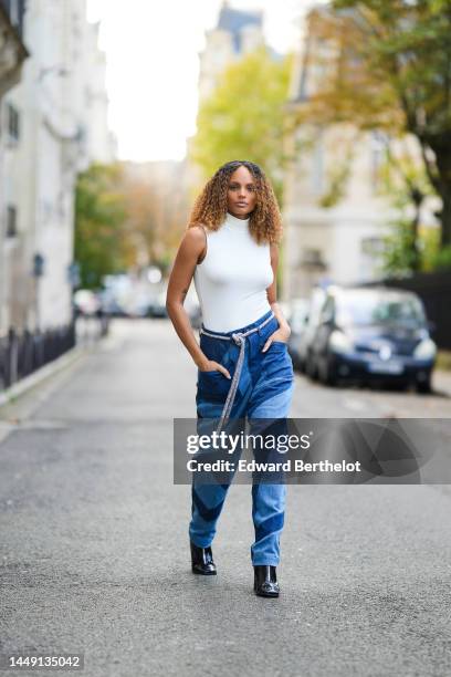 Alicia Aylies wears a white sleeveless / high neck body t-shirt, a gray and black cordon long belt, pale and navy blue embroidered yoke large baggy...