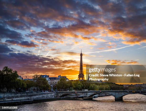 dramatic sunset colors tower above and around the eiffel tower, pont (bridge) des invalides, and surrounding paris skyline as viewed from across the  seine river - sunset with jet contrails stock pictures, royalty-free photos & images