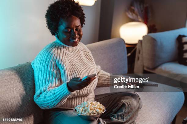 african american woman holding a remote control and smart phone while choosing what to watch on tv - glee tv program stockfoto's en -beelden