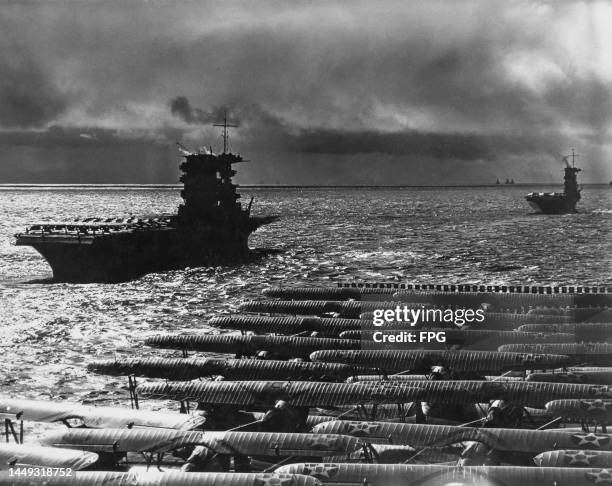 In the foreground, Grumman F3F biplane fighter aircraft sit on the flight deck of the United States Navy aircraft carrier USS Ranger cruising behind...