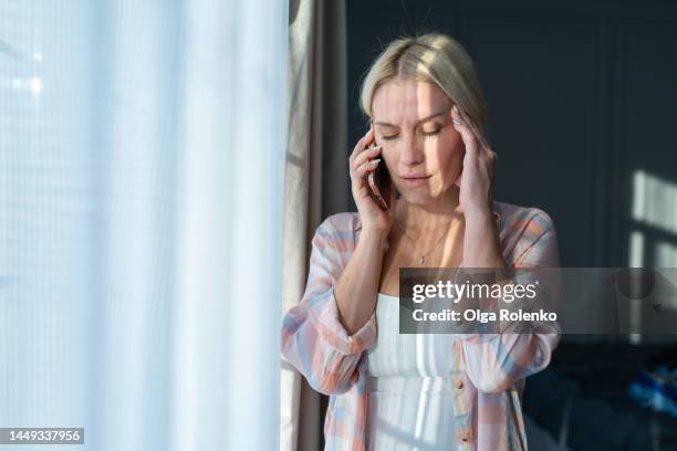 headache and despair. blond woman is on phone, while touching her head near light window - hoofdpijn vrouw stockfoto's en -beelden