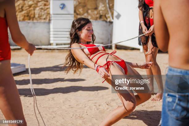 young woman in bikini dancing limbo with friends, bending over backwards and going under the rope - limbo stockfoto's en -beelden