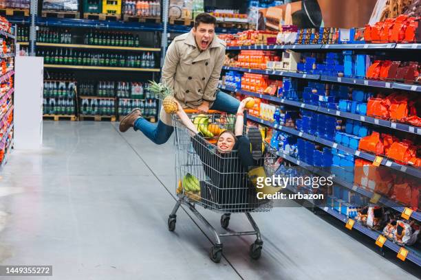 boyfriend pushing his girlfriend in a shopping cart at the grocery store - man pushing cart fun play stock pictures, royalty-free photos & images