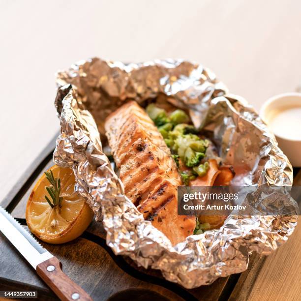 salmon baked in foil with lemon and rosemary and garnished with broccoli. dish is served in foil on wooden board. close-up. soft focus - pavé de saumon photos et images de collection