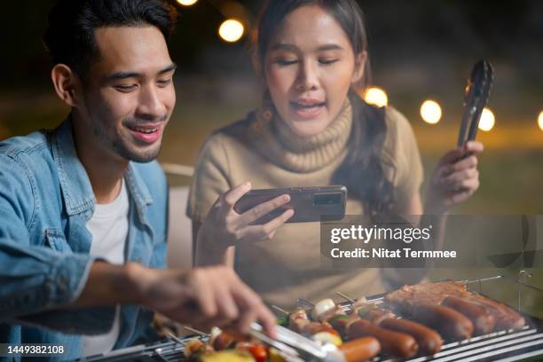 outdoor activities for inclusion of young people. asian males use a tong to grill barbecuing with an open flame grill outdoors and females take a photo or live stream during camping. - daily life in bangkok stock-fotos und bilder