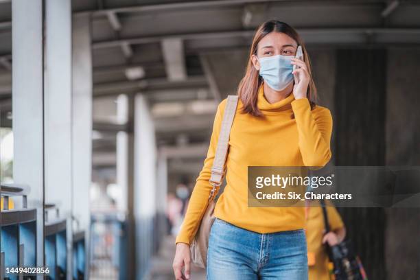 asian businesswoman wearing protective face mask using mobile phone and walking on skywalk at bts sky train railway platform while traveling to work - covid bts stock pictures, royalty-free photos & images