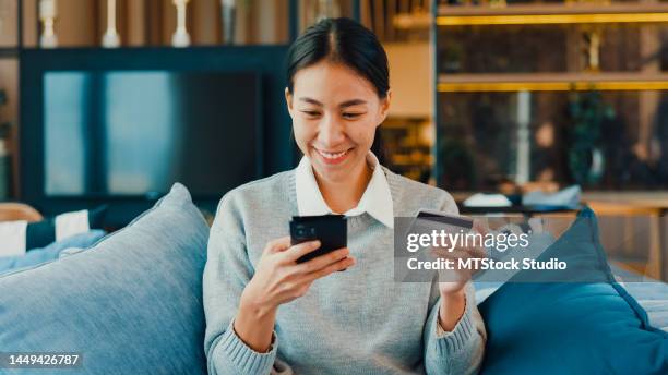cheerful asian young girl wear sweater use smartphone online shopping with credit card on sofa couch in co-working room. - debt free stock pictures, royalty-free photos & images