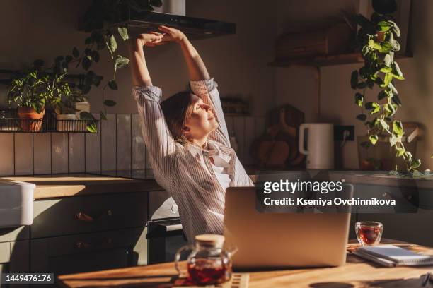 happy relaxed young woman sitting in her kitchen with a laptop in front of her stretching her arms above her head and looking out of the window with a smile - woman front and back stockfoto's en -beelden