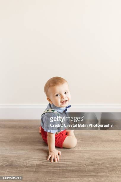 an 11-month-old christmas baby boy with 12 christmas toes, dressed in a bow tie, green christmas tree suspenders, red shorts & a blue short sleeve buttondown shirt sitting on a wooden floor playing, ready for his birthday & christmas in 2022 - bruine ogen stockfoto's en -beelden