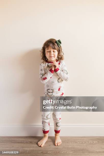 a joyful & hopeful three-year-old toddler girl smiling with her eyes closed, beaning with joy & anticipation while wearing simple neutral christmas pajamas & a dark green hair bow for a cozy christmas at home in december 2022 - young girl white background stock pictures, royalty-free photos & images