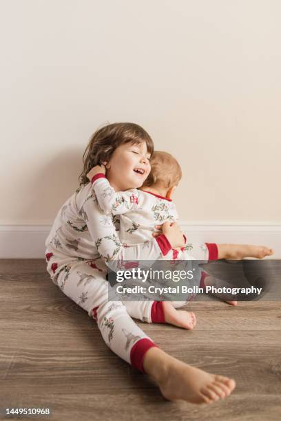 three-year-old toddler girl hugging her 11-month-old baby brother as they wear matching simple christmas pajamas for a cozy christmas season at home in december 2022 - siblings baby stock pictures, royalty-free photos & images