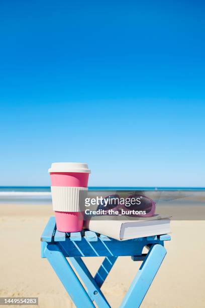 coffee cup, sunglasses and book on a table at beach and sea against sky - buch tisch stock-fotos und bilder