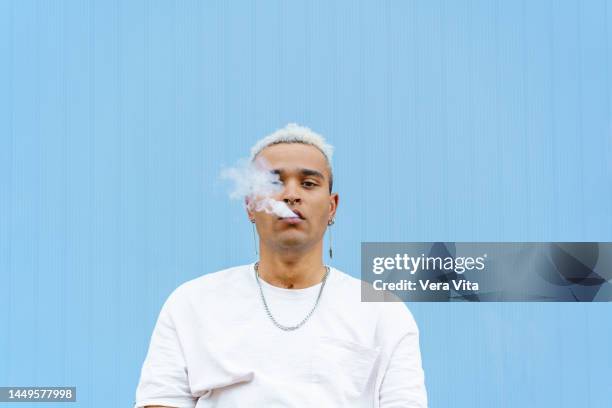 artistic portrait of urban young man with white vape smoke isolated on blue background - vape cigarette stock pictures, royalty-free photos & images